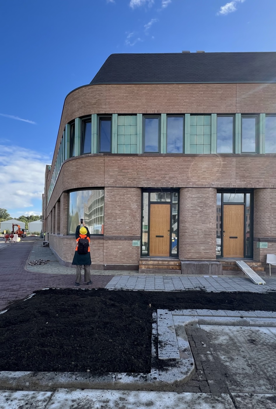 Een deelnemer met bouwhelm en veiligheidshesje fotografeert met een smartphone de gebogen baksteengevel van een nieuw gebouw