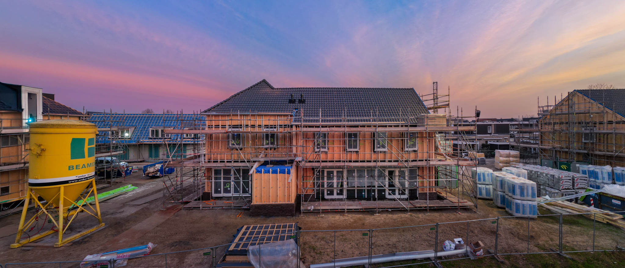 Panorama van een woningbouwproject in aanbouw in de avondschemering, met steigers, een gele silo en een roze en blauwe lucht