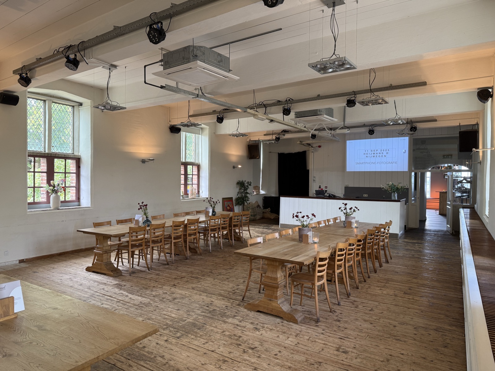 Een zaal met lange houten tafels, klaargezet voor de workshop, met de titelslide Smartphone-fotografie op het scherm